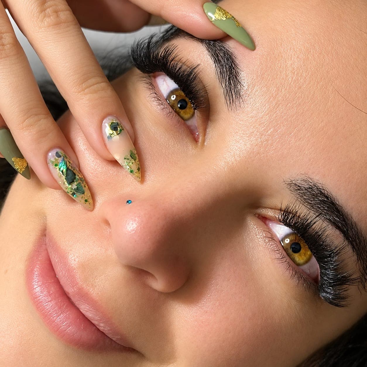 Rosto com extensão de cílios e unhas decoradas em verde e dourado